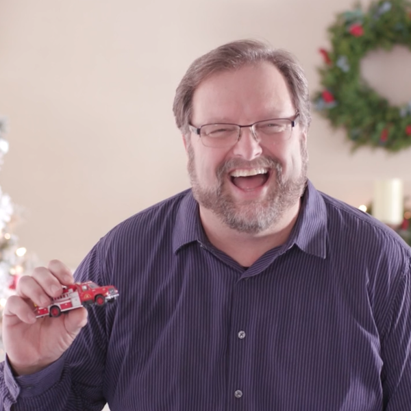 A Keepsake Artist laughs after flubbing a line during a video shoot; he is holding a Keepsake Ornament that he has designed in his right hand; the ornament is shaped like a classic red pickup truck.